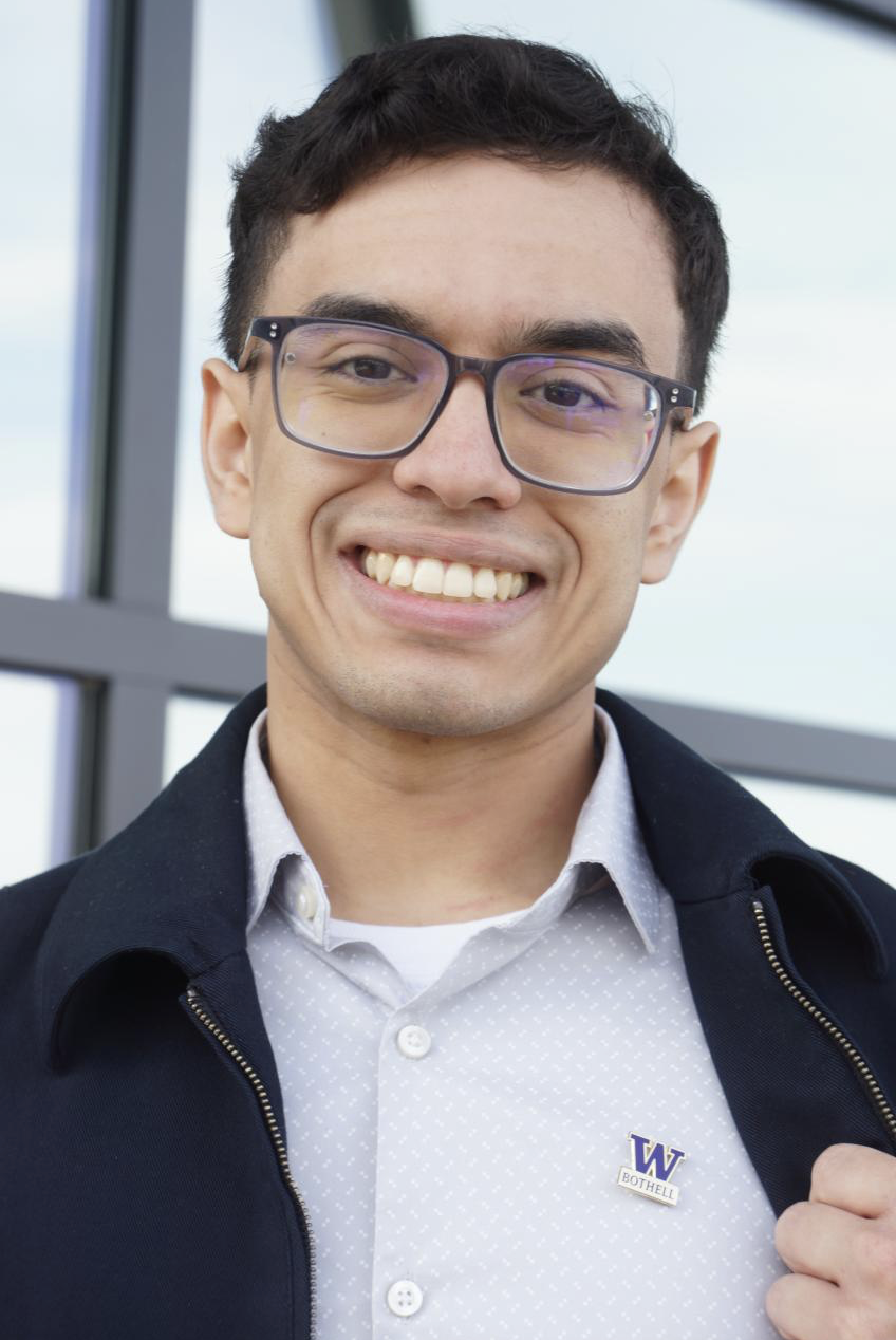 A smiling person with glasses in a blue jacket proudly wearing a UW Bothell pin.